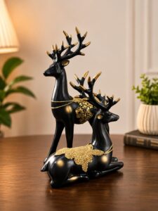 ELEGANT DESIGN: Set of 2 decorative reindeer figurines featuring a sophisticated black finish with luxurious golden accents and ornate floral embellishments, perfect for adding a touch of elegance to any space PREMIUM QUALITY: Crafted from high-quality resin material ensuring durability and long-lasting beauty, these Barahsingha deer sculptures showcase intricate detailing on the antlers and body PERFECT DIMENSIONS: Standing figurine measures 28 cm height x 16 cm length x 5 cm width, whilst the sitting figurine measures 19 cm height x 15 cm length x 4 cm width, ideal for tabletop display VERSATILE DECOR: Suitable for enhancing the aesthetic appeal of living rooms, office desks, mantels, shelves, or as a centrepiece for special occasions and festive celebrations READY TO DISPLAY: Comes as a complete pair with one standing and one sitting deer figurine, featuring detailed golden-tipped antlers and decorative chain accents for an opulent look